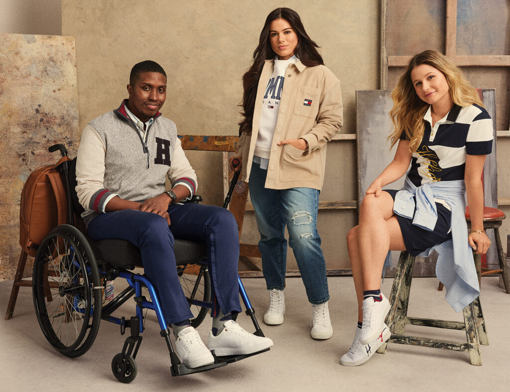 Three models pose for the Fall 2022 Tommy Adaptive campaign; from left to right, a man in a wheelchair wears white sneakers, blue pants and a grey and white zip-up sweater with a large "H" on the chest; a woman using a cane wears white high-top sneakers, denim jeans, a tan jacket with pockets, and a white sweatshirt, both bearing the Tommy Hilfiger logo; a woman seated on a stool wears white sneakers, navy shorts, a light-blue shirt tied around the waist, and a blue-and-white strike polo shirt with the word "Hilfiger" emblazoned in cursive across the body.