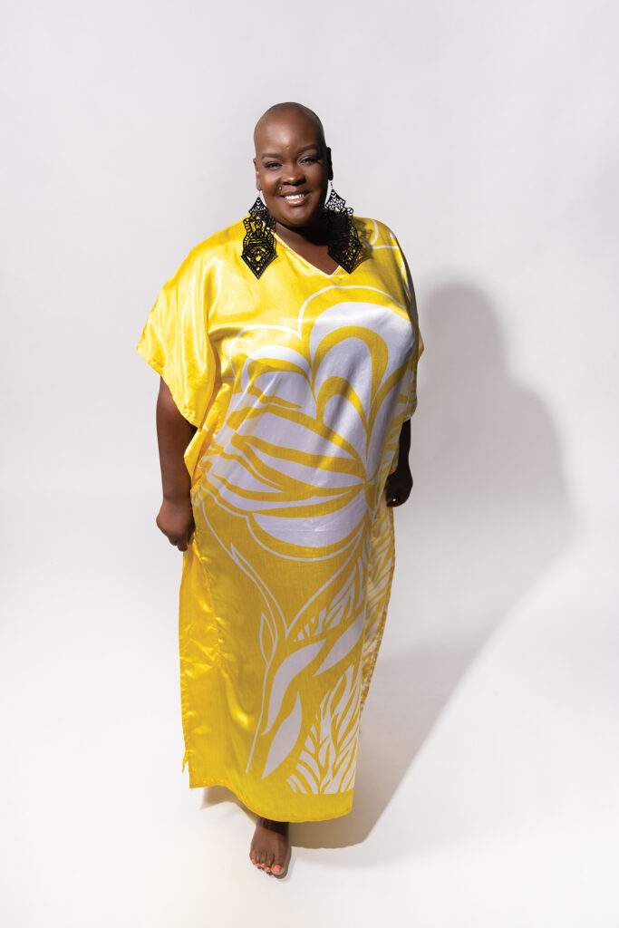 Sonya Renée Taylor stands against an all-white background, hands at her sides and barefoot. She is smiling broadly, wearing long black embroidered earrings, and a full-length yellow satin dress with a large white floral design in the center. 