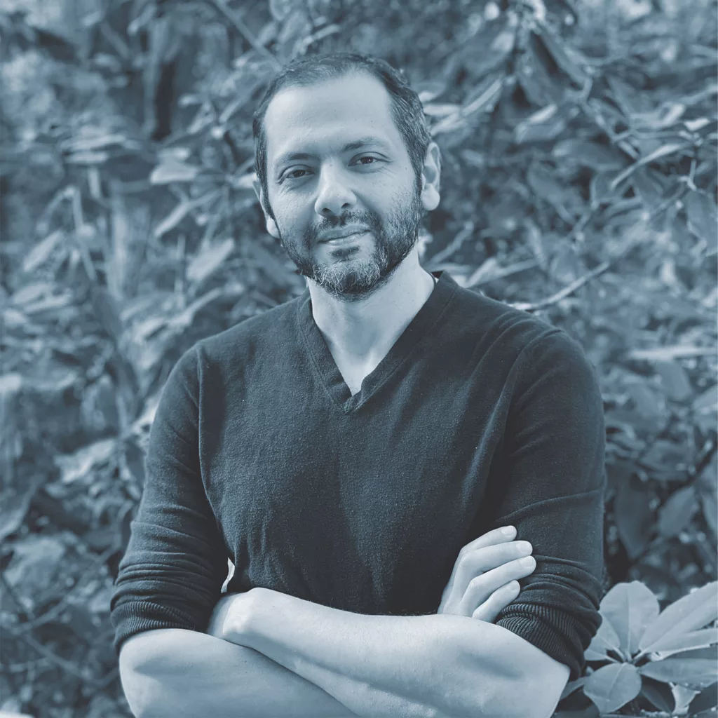A greyscale photo of author Omar El Akkad shows him looking direct to the camera, with his arms crossed over his chest, and foliage in the background.