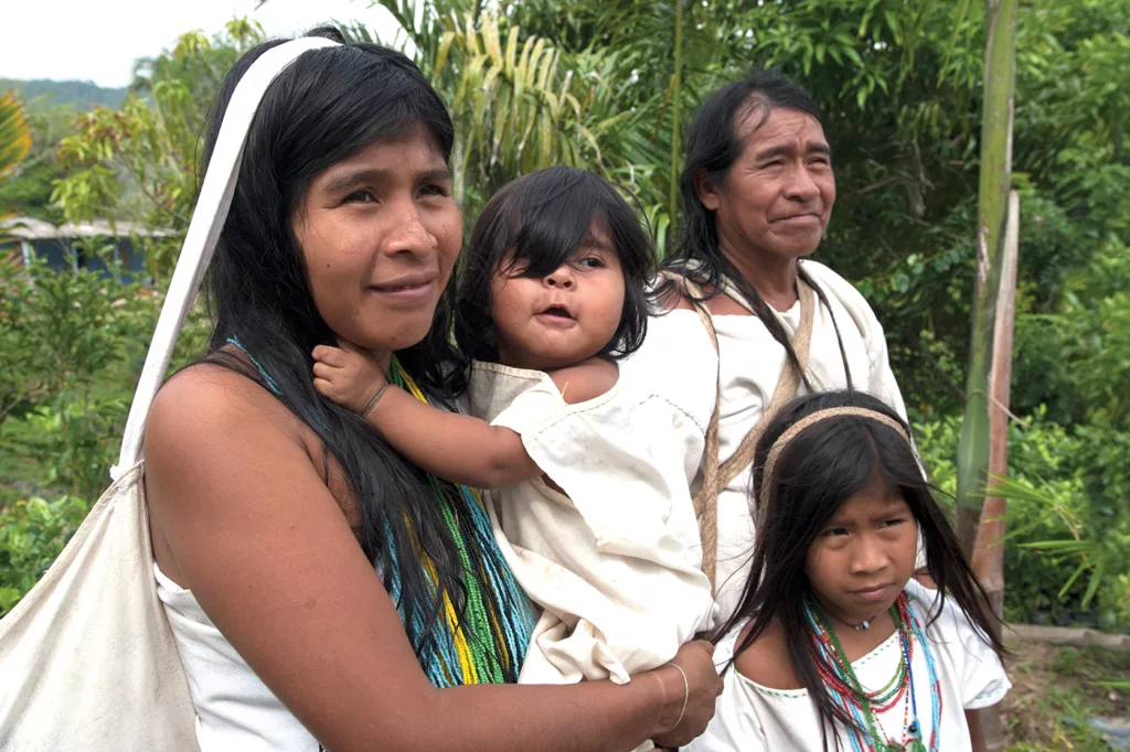This Kogui family includes a mother, father, and two children, all dressed in white. The mother and older child are also wearing brightly colored necklaces made from beads. 