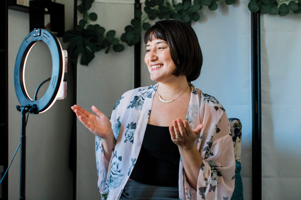 Bex Mui smiles into her phone, mounted on a ring light. She is seated, wearing a floral blouse and black tank top, and her hands are raised to mid-chest level, palms up.  