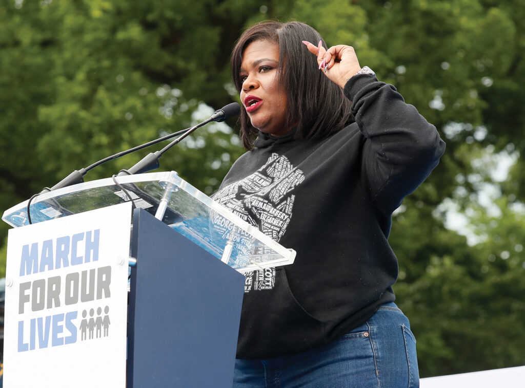Rep. Cori Bush, wearing a black hoodie with a fist on it, stands at a March for Our Lives podium. 