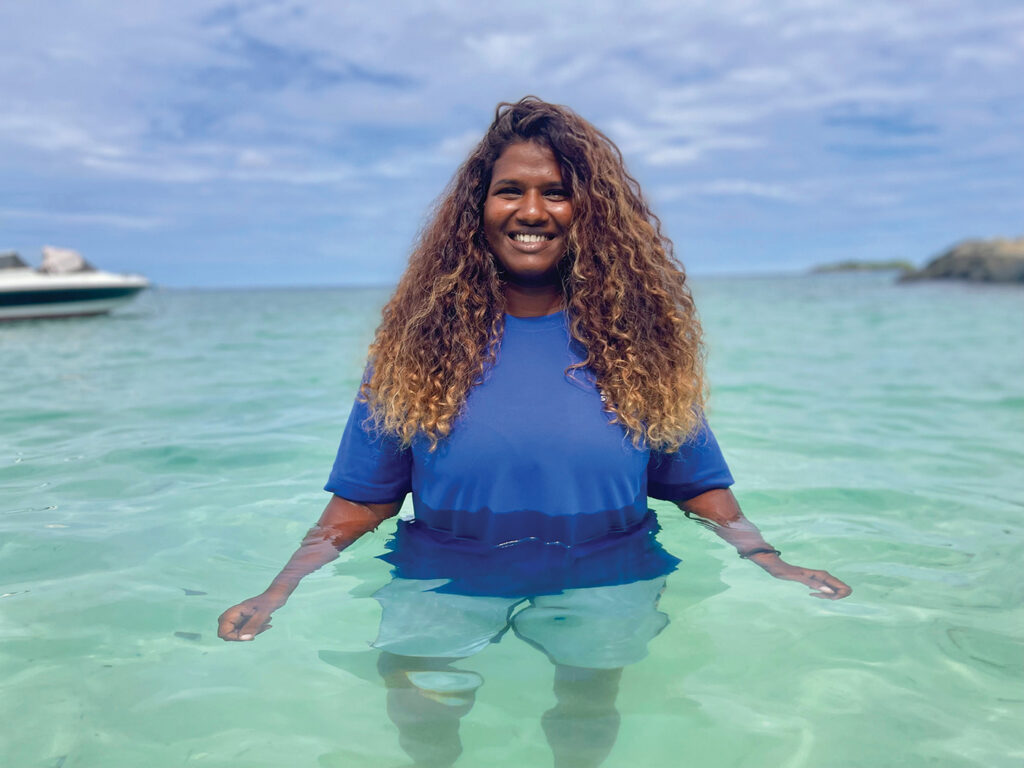 Aminath Zoona, stands beaming in waist-high turquoise ocean water. She has long wavy hair and wears a blue shirt and shorts.