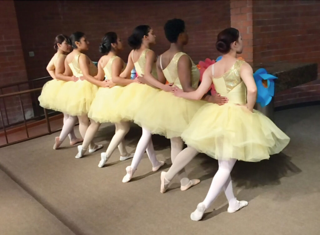 Six young woman from The Swan Within in mid dance. They are dressed identically in yellow tutus and have different skin colors.