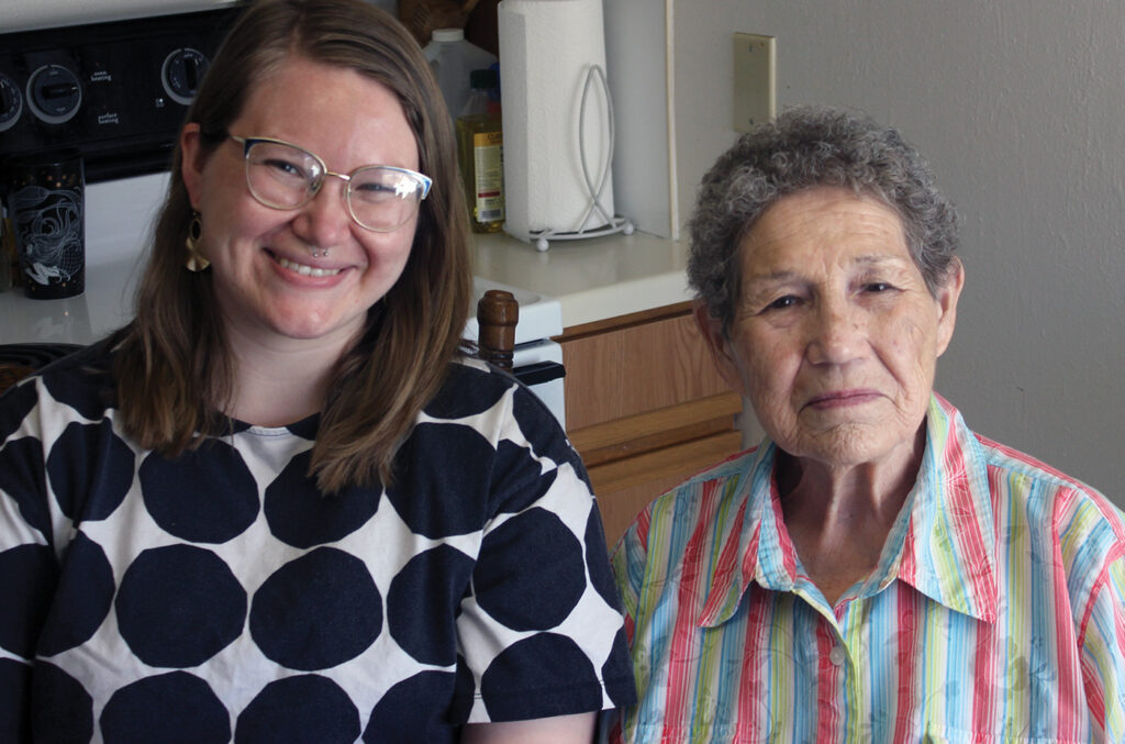 A photograph from 2022 of author Abaki Beck, smiling in a black and white graphic dress, next to her grandmother, Angeline Wall, an older woman in a striped shirt. They are seated at a kitchen table. 