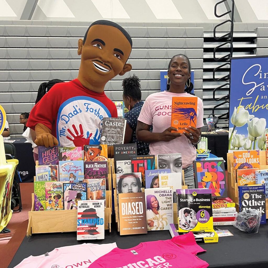 A photograph of Iesha Malone behind a Rose Cafe table at the Wonder Literary Festival in Chicago's South Side. The table has a mix of children's and adult books. Iesha holds up the book Nightcrawling by Leila Mottley and smiles. 