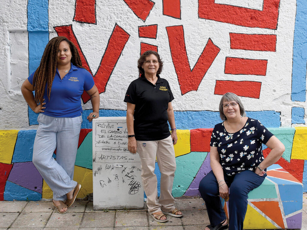 Three women look on in front of a colorful wall, on which the words Rio Vive are painted. The women are Mónica Ponce-Caballero, Mercedes Rivera Morales, and Ana Luisa Baca, who have all had leadership positions at CAUCE. 