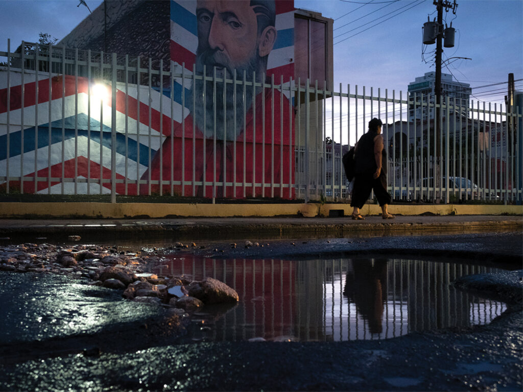 A picture of dusk on Calle Cerre, revealing a large pothole filled with water. A large mural is in the background, and its subject, a man with a beard, looks down on the street. 