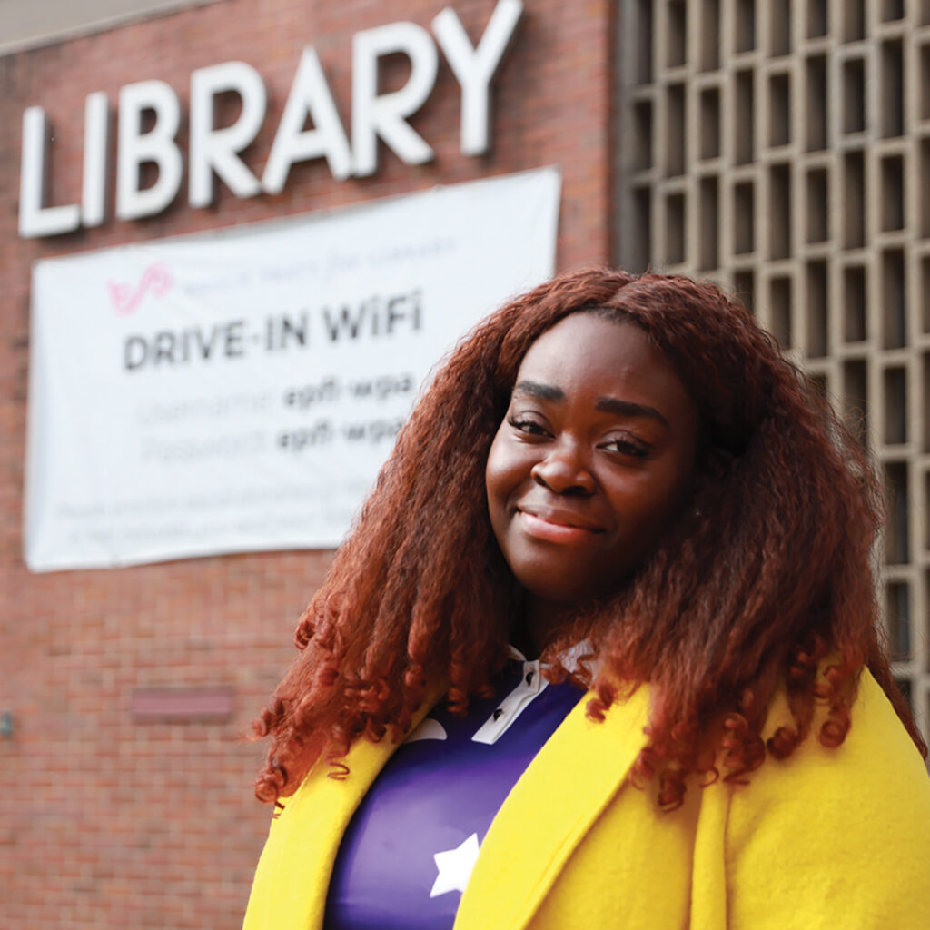 Storybook Maze, a black women with red-hued hair wearing a bright yellow jacket, smiles at the camera outside a public library in Baltimore