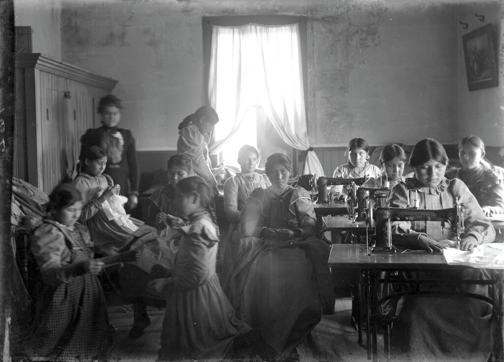 A black and white photo depicting an interior at the Cut Bank Boarding School. 11 female Native "students," dressed in long-sleeved dresses, sit at sewing machines or do hand-mending. Some are looking at the camera and some are not. 