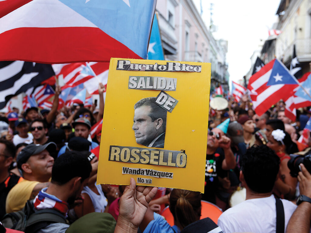 A photograph from August 2, 2019 of a crowded outdoor street packed with people waving Puerto Rican flags. In the foreground is a handmade yellow sign that reads "Puerto Rico salida de Rossello, Wanda Vasquez no va" with a picture of Rossello on it. 