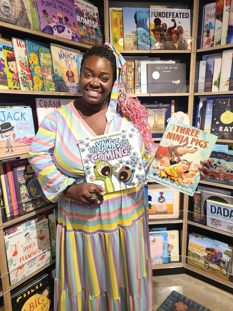 Maze Storybook, a black woman wearing a colorful, striped dress, smiles as she shows off two children's books from within an independent bookstore in California. 