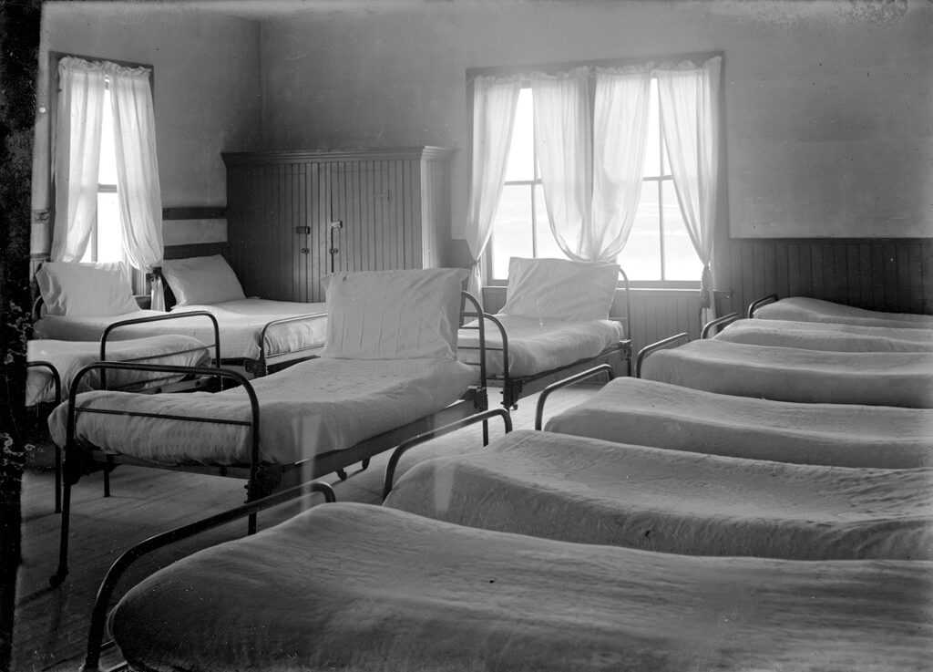 A black and white photo from the Cut Bank Boarding School of a bedroom with at least 14 beds in it. The beds are empty and made with white sheets. 