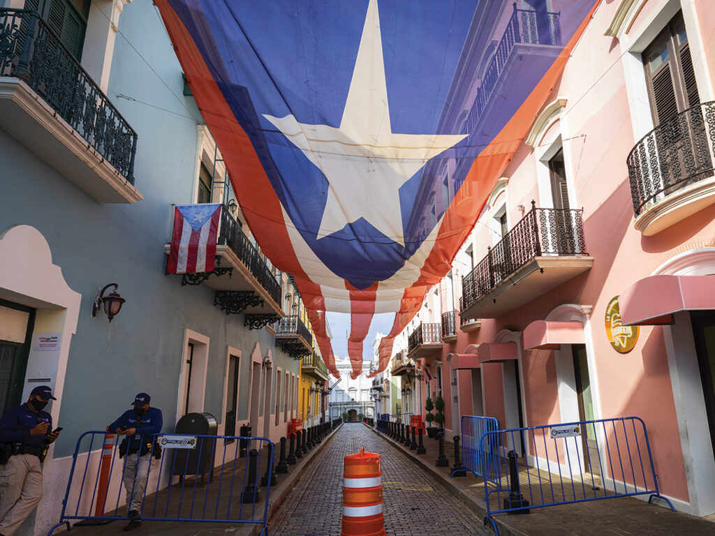 A photograph of an empty Puerto Rican street. A large Puerto Rican flag flows down the top of the street, and two masked security guards look on. 