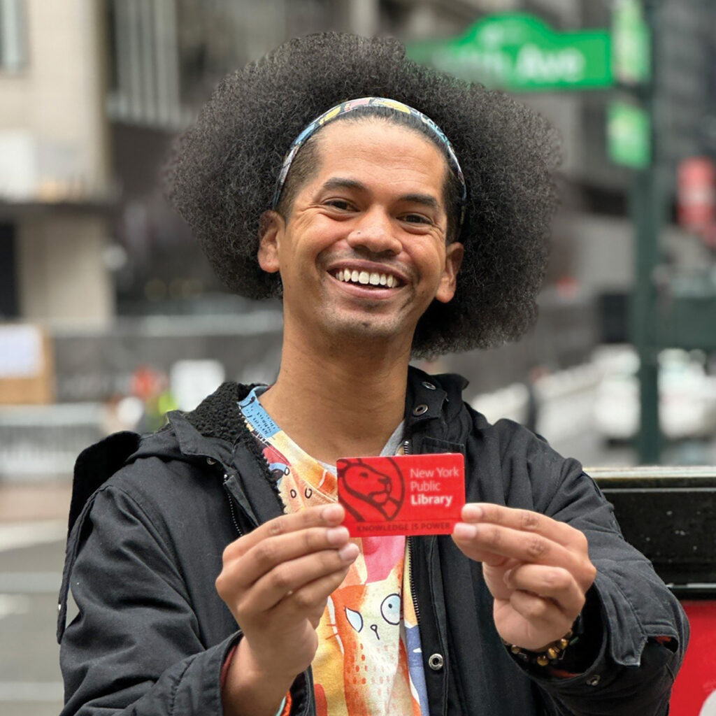 Mychal Threets, a biracial young man, smiles as he holds up a red New York Public Library card. 