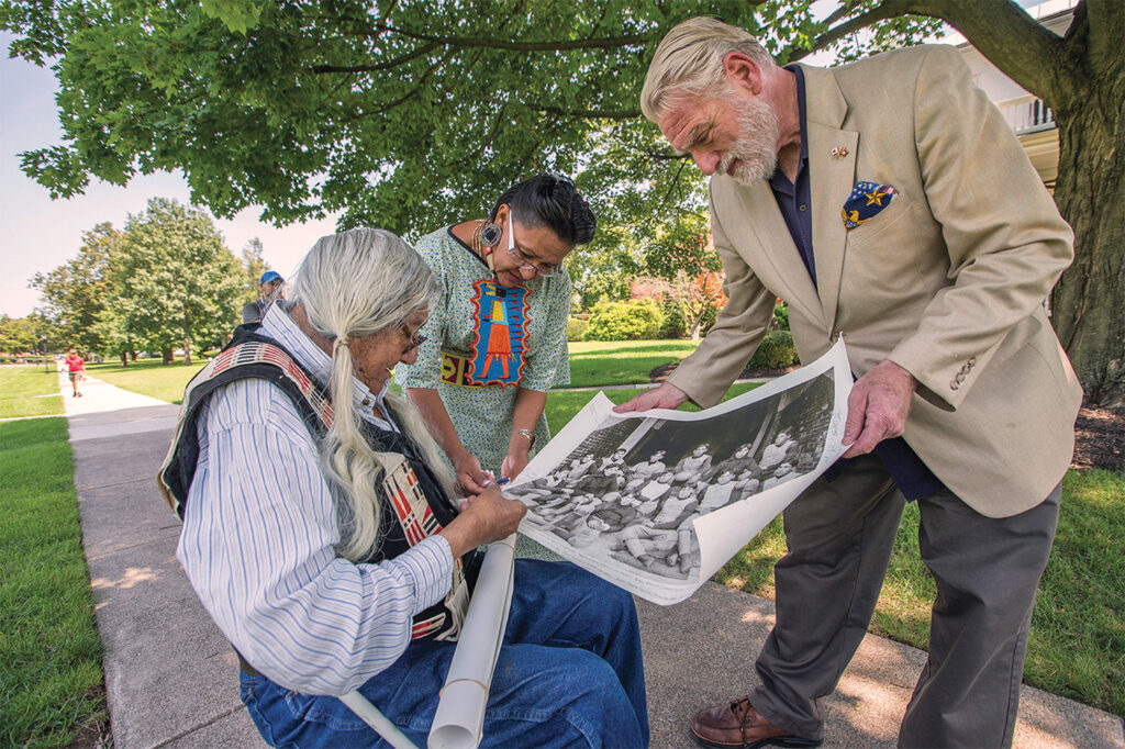 A photograph of Mark Soldier Wolf, an older, seated Native man with long white hair, and his daughter, Yufna Soldier Wolf, who stands next to him. They are looking at a print out of a black and white photo of a group of children from Carlisle Barracks, a boarding school located in Carlisle, Pennsylvania.