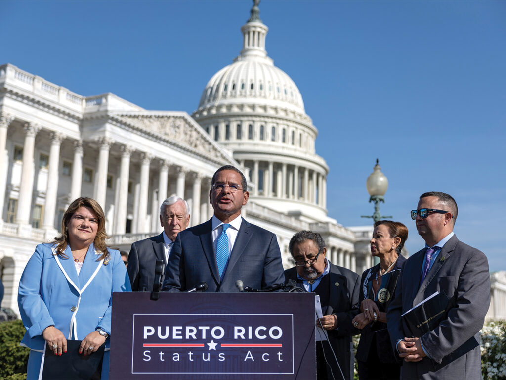 A photograph from April 2023 showing Puerto Rico Governor Pedro Pierluisi speaking at a podium labelled with the words Puerto Rico Status Act. He is outside, and the Capitol building is behind him. He is flanked by three men and two women who look on while he speaks. 