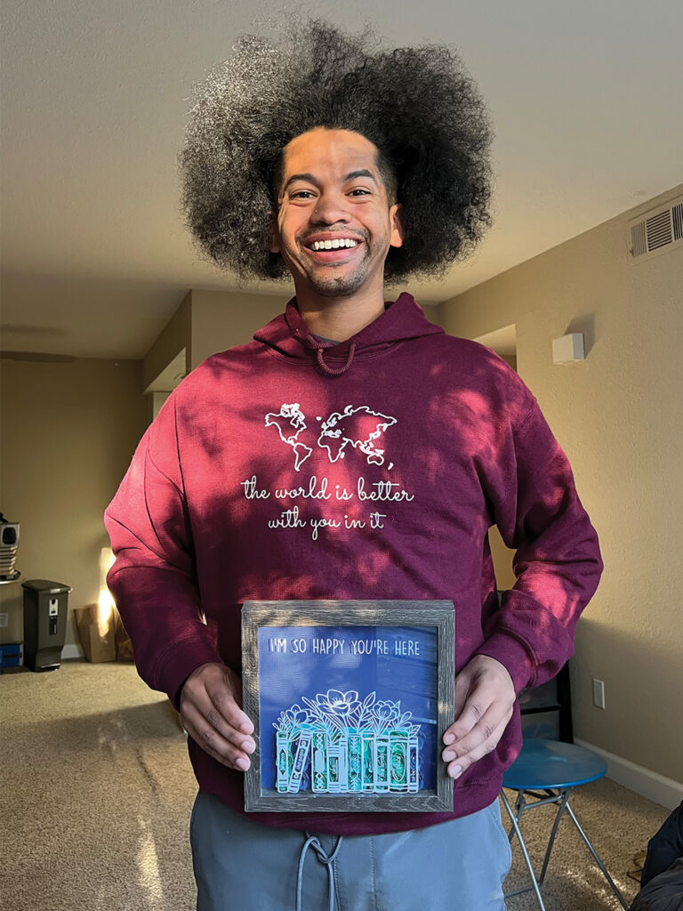 Mychal Threets, a biracial young man, wears a giant smile and a maroon hoodie that reads "The world is better with you in it." He is holding a framed photo that reads "I'm so happy you're here" with an illustration of books and flowers. 