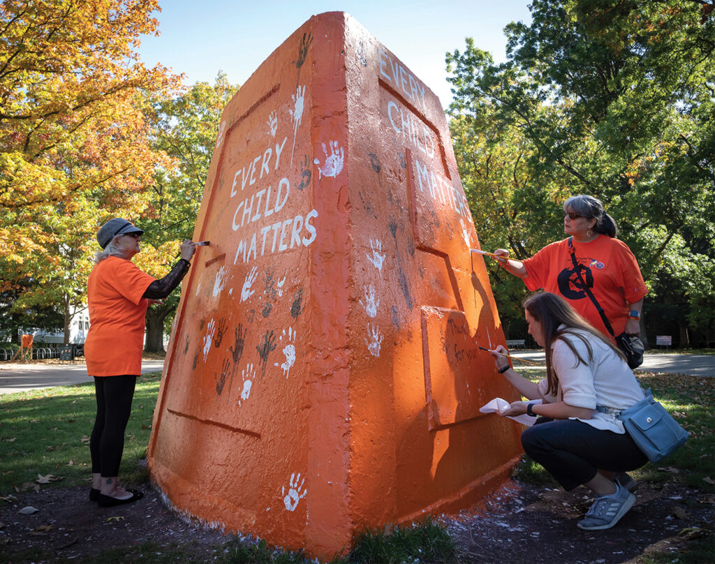 Three women, two in orange shirts, surround a large, orange-painted concrete structure. "Every child matters" is painted on it in white, which became a message of solidarity in Canada after the remains of children were found in boarding school grounds. 
