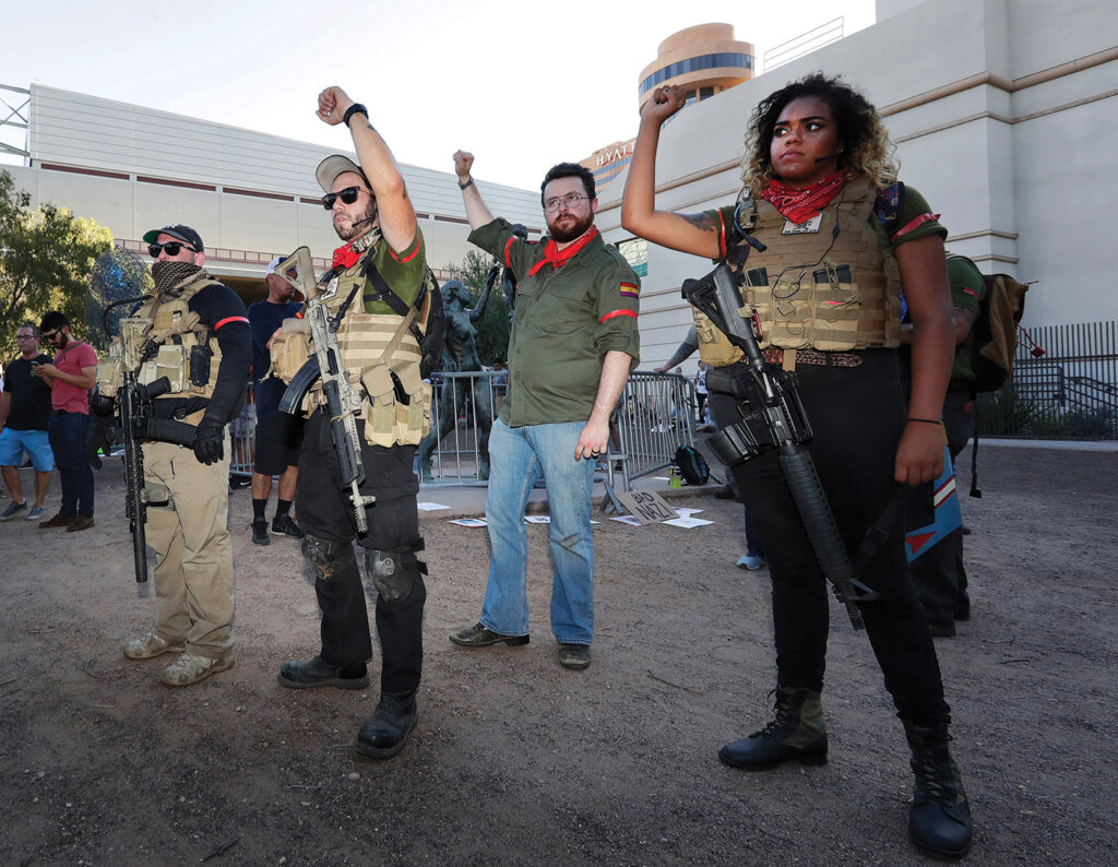 A photograph with four people, all with red armbands, in the foreground, protesting Trump on Aug. 22, 2017. Three on the left are white men, and the person on the right is a brown-skinned woman. Three of the people have automatic weapons, including the woman.