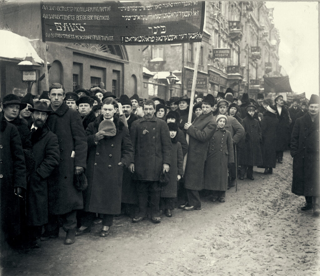 A black and white photograph of the Jewish Labour Bund in Moscow, 1917. A large group of people, mostly men, are shown, in winter coats and hats. They are holding a placard in both Russian and Yiddish, that reads, in part, “Hail the Russian Social Democratic Workers Party! Hail the General Jewish Workers Union!"