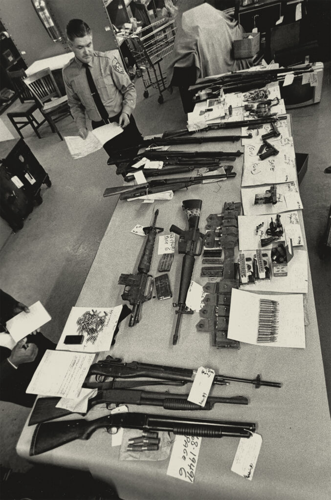 A black and white photograph from 1968 shows a white police officer looking over two tables on which are a number of guns and ammunition confiscated from the Oakland Black Panthers.