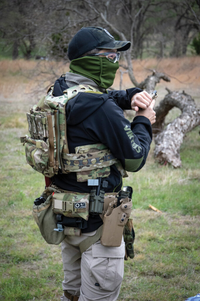 An Asian American person wears tactical garments, a belt holding many tools, a backpack, and holds a gun. Their face and hair are covered. They are leading a training for Yellow Peril Tactical.