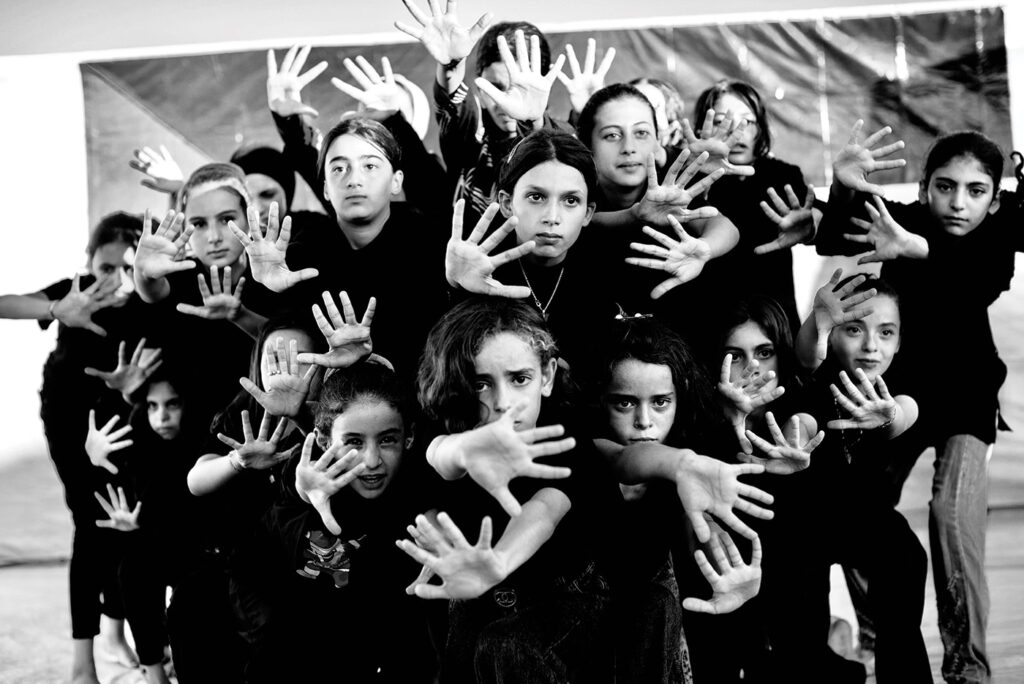 A black and white photo of the Al Fursan dance troupe. 18 young girls wearing black hold out their hands, creating a striking, artistic photo.
