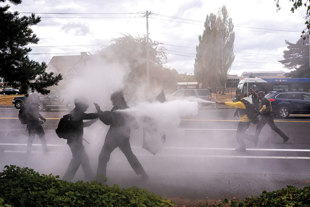 A picture of far-left and far-right activists clashing in a street in Portland, Oregon, on Aug. 22, 2021. There is white smoke or gas among the people fighting, and cars and a city bus are seen behind them.