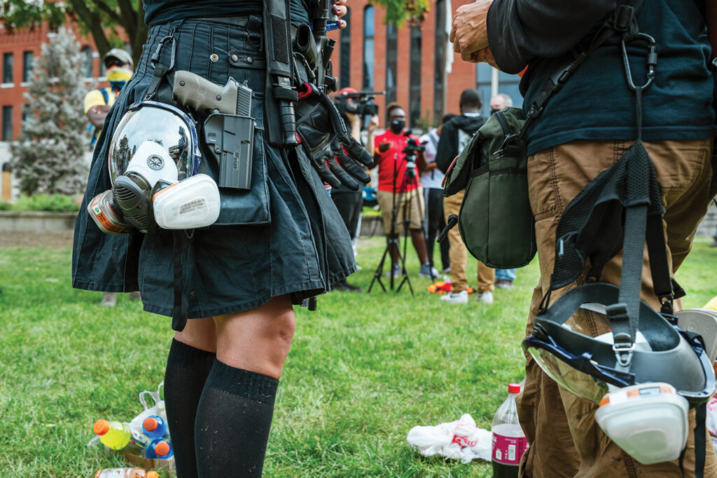 A photograph from protesters in Louisville, KY, in 2020 protesting the judicial responses to Breonna Taylor's killing. The person on the left, a woman, is strapped with multiple weapons, including a gun and a knife. She also has a gas mask and gloves. The person on the right, a man, has a gas mask. Both appear to be white. In the background, both black and white protesters gather near a digital camera.