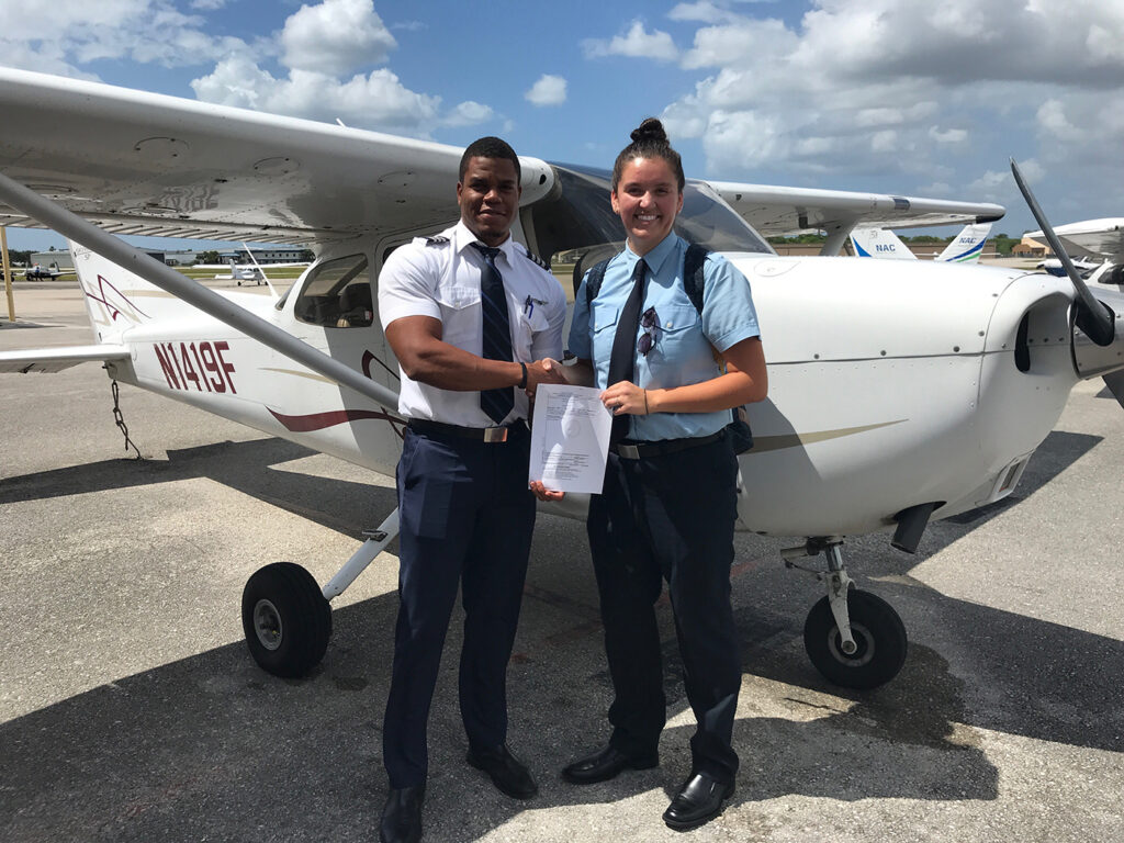 Shawnee Real Bird, a young Native woman, smiles broadly and holds a sheet of paper, her pilot's exam. She is standing next another pilot, who is black—her flight instructor.  