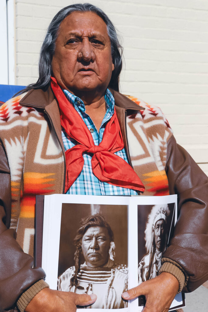 An older Native man with long hair, a brown leather jacket, and red bandana holds a hardback book open. The book is open on a sepia-toned photo of a Native man from the 1800s, his ancestor. 