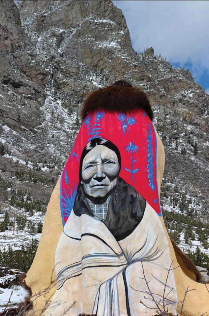 A figure stands facing a majestic, large, rock formation in the background. They are wearing a buffalo robe with a tallow painting by Miah Chalfant—a black-and-white portrait of a tribe matriarch wrapped in a white blanket. Behind her is a red background with blue herbs and flowers decorating it. 