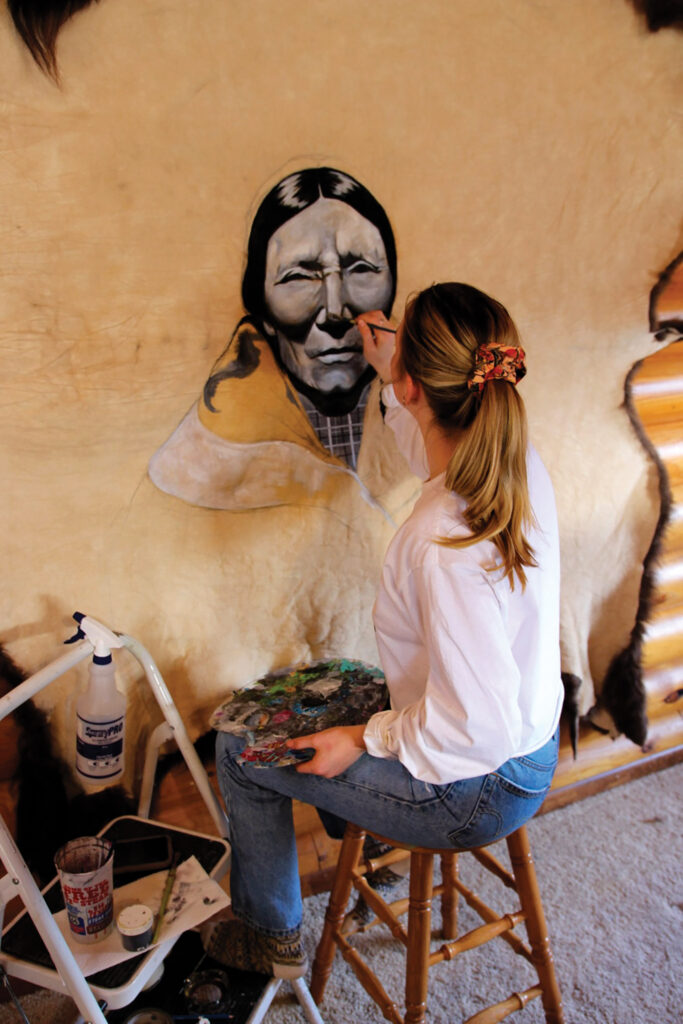 A picture from the back of Mia Chalfant painting on hid. She has her hair pulled back in a ponytail and holds a painter's palette of her natural tallow pigments.