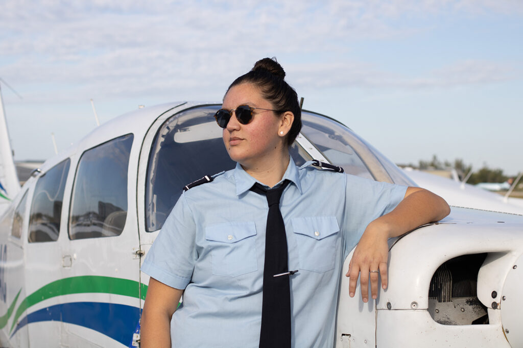 Shawnee Real Bird, a young Native woman, stands confidently in front of a small airplane, resting her arm on the plane's nose. She is wearing her hair in an upright bun, and sports aviator glasses and a pilot's outfit of a blue collared shirt and black tie.